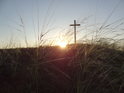 Velký ocelový kříž na vrcholu Radobýlu při zvedajícím se ranním Slunci za suchomilonými travinami.